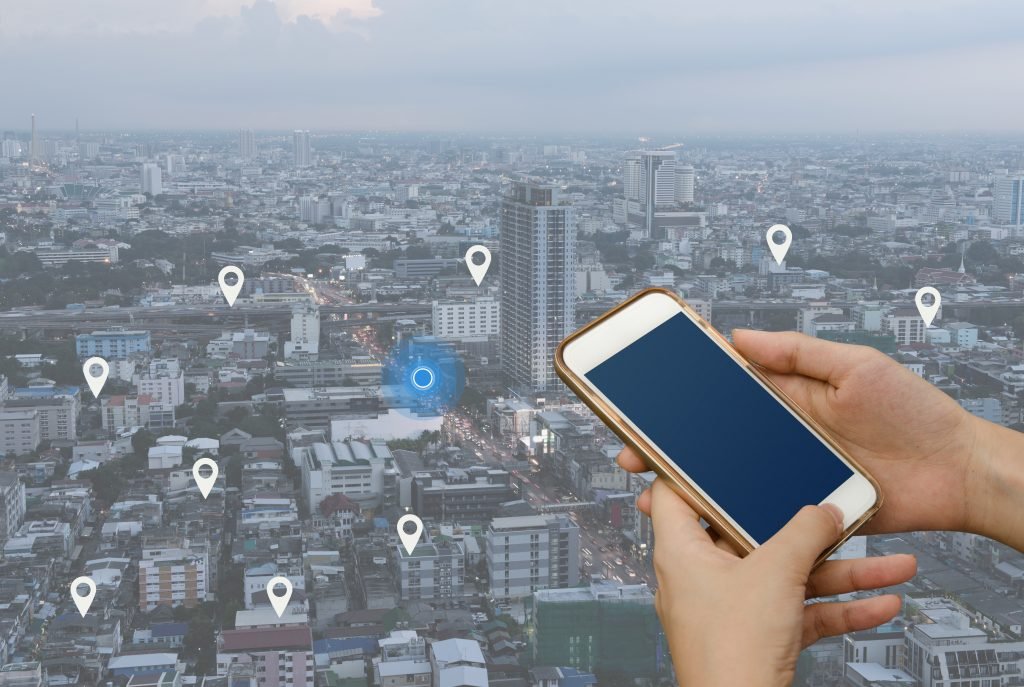 Woman hand using mobile smartphone searching location on map and pin above blue tone city scape and network connection, internet of things, satellite navigation system app, smart journey concept