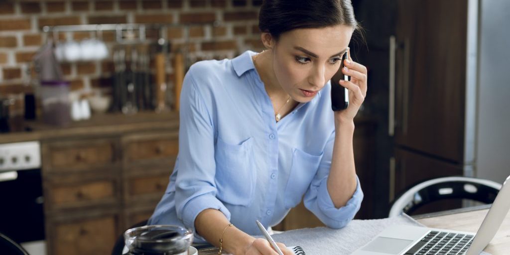 Concentrated young woman using laptop and smartphone while making notes